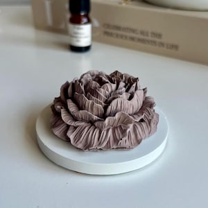 May include: A taupe-coloured, sculpted flower ornament with intricate, layered petal details, resting on a smooth, circular white base. The flower features a dense, ruffled texture throughout its form, creating a realistic botanical appearance.