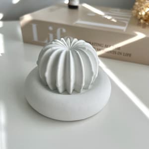 May include: A white, cactus-shaped ornament rests on a white, circular base. The cactus has vertical ridges, and the base is smooth. The objects are on a white surface, with a book and other items in the background.