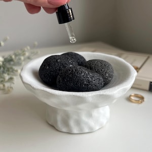 May include: A white ceramic pedestal bowl with a textured, uneven rim and base, containing several porous black lava stones. A glass dropper with a black cap is positioned above the bowl, ready to dispense liquid onto the dark volcanic rocks.