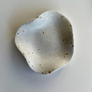 May include: A small, irregularly shaped dish with a light grey, speckled surface. The material features embedded, multi-coloured flecks and small, scattered indentations throughout the concave interior, creating a textured, stone-like appearance.