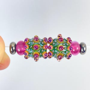 May include: A handmade glass bead with a central section of green and yellow, adorned with purple and orange accents. The bead is flanked by two pink beads and two silver-colored end caps. The bead is strung on a thin, clear cord.