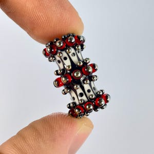 Puede incluir: Primer plano de una cuenta decorativa. La cuenta presenta un patrón en blanco y negro con detalles en rojo y plata. La cuenta se sostiene entre dos dedos sobre un fondo blanco.