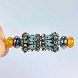 以下が含まれることがあります： さまざまな色のガラスビーズの連なり。中央のビーズは、金と銀のアクセントが付いた水色です。両側のビーズは銀色と琥珀色です。ビーズは透明で細いコードに通されています。