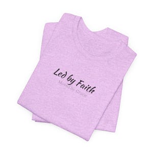 May include: A folded light purple t-shirt with the text "Led by Faith, Moved by Grace" in black script. The shirt is made of a soft material and is neatly folded, showcasing the design on the front.
