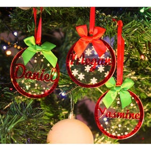 Può includere: Tre decorazioni natalizie trasparenti con bordi rossi e nomi: Daniel, Megan e Yasmine. Ogni ornamento ha un nastro rosso e un fiocco verde. Gli ornamenti sono appesi a un albero di Natale con luci.