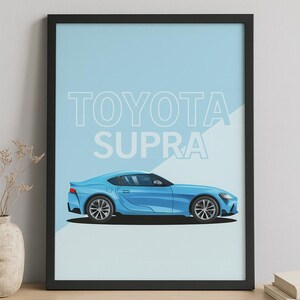 May include: A framed print showing a light blue Toyota Supra car illustration. The car is in profile against a light blue backdrop, with the words "TOYOTA SUPRA" in white above. The frame is black, and the print is on display.