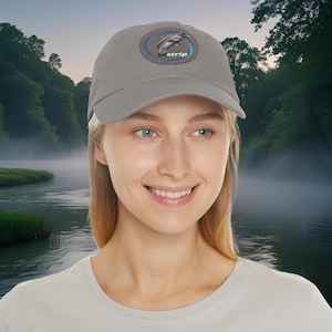 May include: A gray baseball cap with a curved brim. The cap features a circular patch with a fish leaping out of the water, surrounded by a blue ring and the word "strip". The background shows a river and trees.