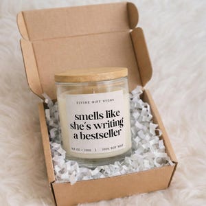 Peut inclure: Bougie en pot de verre avec couvercle en bois, étiquetée "smells like she's writing a bestseller". La bougie est dans une boîte en carton avec du papier de remplissage blanc. La bougie pèse 130g et est faite à 100% de cire de soja.