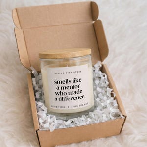 Op de afbeelding: Een kaars in helder glas met lichte was en een houten deksel, geplaatst in een bruine kartonnen doos gevuld met witte papieren snippers. Het etiket vermeldt: DIVINE GIFT STORE, smells like a mentor who made a difference, 130g, 100% sojawas.