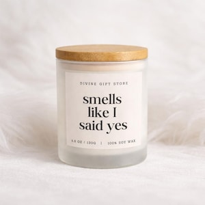 Könnte beinhalten: Eine Kerze aus mattiertem Glas mit einem Holzdeckel. Auf dem Etikett steht "smells like I said yes" in einer fetten, modernen Schriftart. Die Kerze wiegt 130g und besteht aus 100% Sojawachs. Der Markenname "DIVINE GIFT STORE" ist darüber gedruckt.