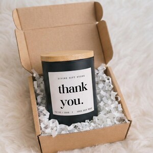 Op de afbeelding: Een zwarte kaars in een cilindrische houder met een lichthouten deksel, geplaatst in een kartonnen geschenkdoos met wit papieropvulmateriaal. Het witte etiket vermeldt Divine Gift Store, thank you., 130g, 100% sojawas in zwarte letters.
