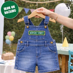 Op de afbeelding: Blauwe denim overall shorts met de tekst "KAYCEE STREET" op een patch. De shorts hangen aan een houten hanger. De afbeelding bevat ook een groene cirkel met de tekst "IRON ON BACKING".