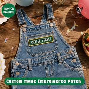 May include: Blue denim overalls with a rectangular embroidered patch that reads "BRYLEE STREET" in white letters on a green background. The overalls have metal buttons and adjustable straps. The text "IRON ON BACKING" and "Custom made Embroidered patch" are also visible.