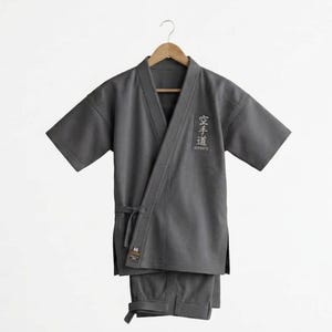 May include: A dark gray martial arts uniform consisting of a short-sleeved jacket and matching pants hanging on a wooden hanger. The jacket features a wrap-around front with a tie closure and white Japanese kanji characters embroidered on the chest.