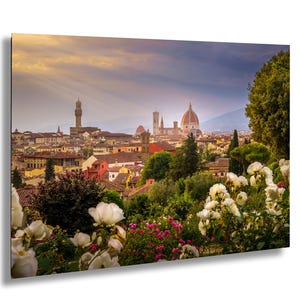 Puede incluir: Una vista panorámica de Florencia, Italia, con el Duomo y otros edificios históricos bajo un cielo dramático. El primer plano muestra flores blancas y rosas, con exuberante vegetación. La imagen es una representación vibrante de la ciudad.