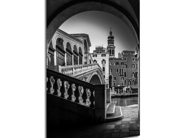 May include: Black and white photograph of a Venetian scene, framed by an archway. The Rialto Bridge is visible, with buildings and a canal in the background. The image is a study in light and shadow.