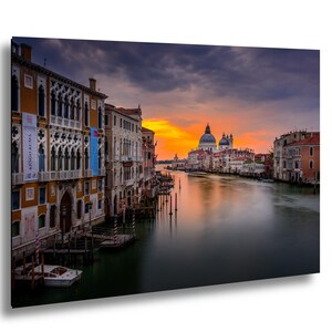 May include: A framed canvas print of Venice, Italy, featuring buildings along a canal at sunset. The sky transitions from orange to dark blue, reflecting in the water. Buildings have arched windows and ornate details.