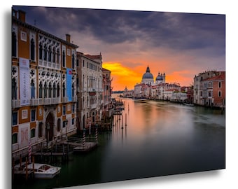 May include: A framed canvas print of Venice, Italy, featuring buildings along a canal at sunset. The sky transitions from orange to dark blue, reflecting in the water. Buildings have arched windows and ornate details.