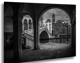 May include: Black and white photograph of a Venetian scene. The Rialto Bridge is visible through an archway, with buildings and a canal in the background. A street lamp stands on the right, and a staircase is on the left.