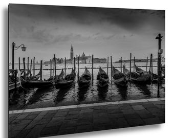 May include: A monochrome photograph of gondolas moored in Venice, Italy. The image shows several gondolas tied to wooden poles in the water, with a cityscape and overcast sky in the background. The scene is a classic representation of Venetian architecture.