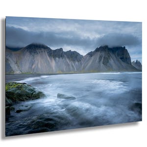 May include: A landscape photograph of a beach with a mountain range in the background. The sky is overcast with dark clouds. The water is a blurred, milky blue, and the sand is dark. A person is visible on the beach.
