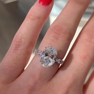 May include: A diamond engagement ring with an oval-cut center stone and smaller accent stones. The ring is set on a delicate band and is displayed on a hand with red nail polish. The text "Brilliant Jewels Art" is visible.