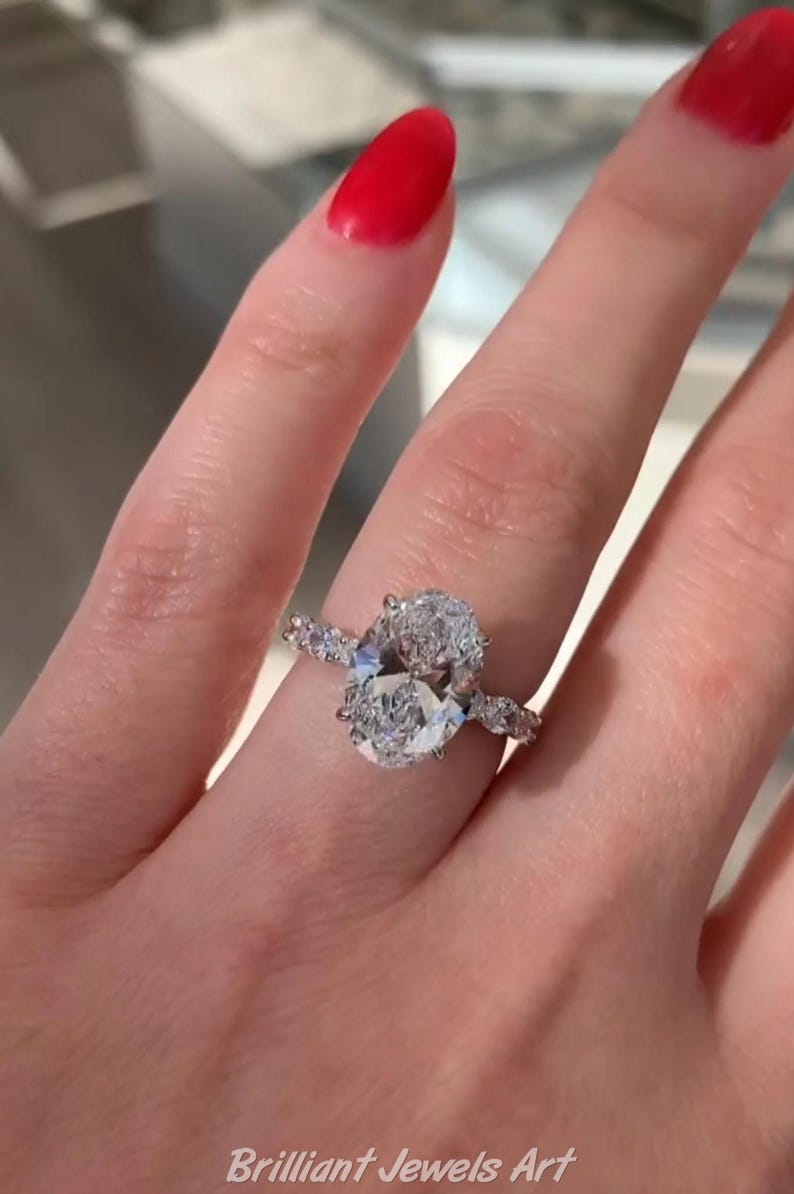 May include: A diamond engagement ring with an oval-cut center stone and smaller accent stones. The ring is set on a delicate band and is displayed on a hand with red nail polish. The text "Brilliant Jewels Art" is visible.