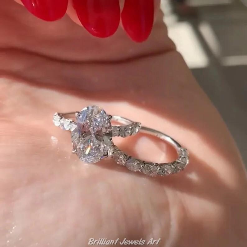 May include: Two silver rings with clear gemstones. One ring features a large oval-cut stone, while the other has smaller stones set around the band. The rings are displayed on a light pink surface. The text "Brilliant Jewels Art" is at the bottom.