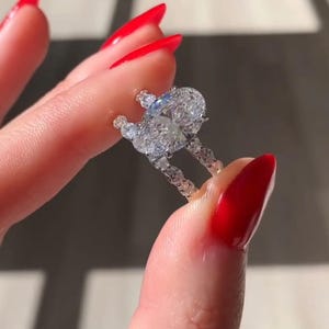 May include: A diamond ring with a large oval-cut center stone and smaller accent stones. The ring is held between two fingers with red nail polish. The ring is set in a delicate band.