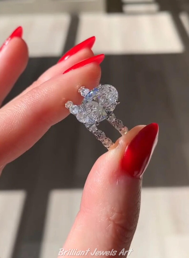 May include: A diamond ring with a large oval-cut center stone and smaller accent stones. The ring is held between two fingers with red nail polish. The ring is set in a delicate band.