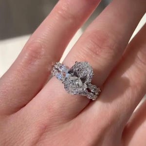 May include: A diamond engagement ring with an oval-cut center stone and a pav&eacute; band. The ring is set on a finger with red nail polish. The text "Brilliant Jewels Art" is visible at the bottom of the image.
