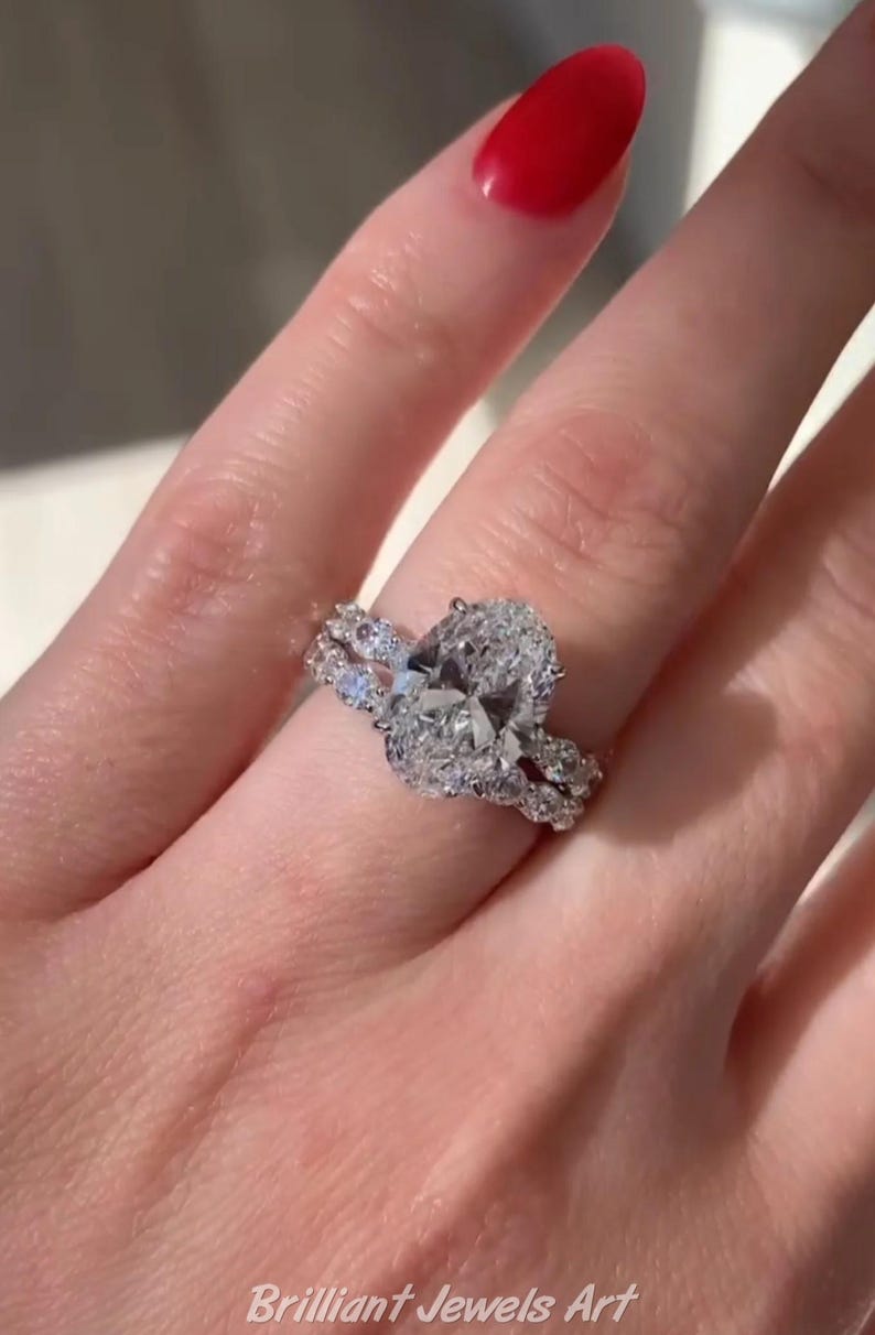 May include: A diamond engagement ring with an oval-cut center stone and a pav&eacute; band. The ring is set on a finger with red nail polish. The text "Brilliant Jewels Art" is visible at the bottom of the image.