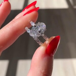 May include: A diamond ring with a large oval-cut center stone and smaller accent stones. The ring is held between fingers with red nail polish. The ring is set in a white metal, likely platinum or white gold. The ring is a piece of fine jewelry.