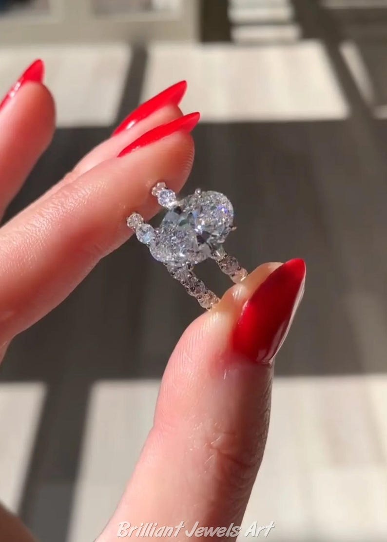 May include: A diamond ring with a large oval-cut center stone and smaller accent stones. The ring is held between fingers with red nail polish. The ring is set in a white metal, likely platinum or white gold. The ring is a piece of fine jewelry.
