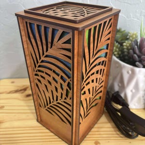 Puede incluir: Una lámpara de madera con forma cuadrada y un diseño de hojas de palmera cortado en los lados. La lámpara es de color marrón claro y tiene una parte superior cuadrada con un patrón de hojas similar. El interior de la lámpara es visible a través de los recortes, revelando colores azules y verdes.