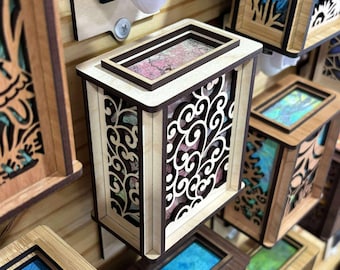 May include: Wooden night lights with intricate cut-out designs. The night lights have a rectangular shape with a top frame and a decorative front panel. The colors include light and dark brown wood, with some featuring colorful inserts. The night lights are plugged into a wall outlet.