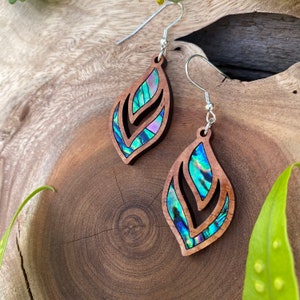 Peut inclure: Une paire de boucles d'oreilles en bois avec un motif de feuille. Les feuilles sont incrustées de nacre d'ormeau irisée, créant un magnifique motif bleu et vert. Les boucles d'oreilles sont attachées à des crochets argentés.