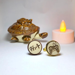 Può includere: Due gemelli in legno, uno con le iniziali "HA" e l'altro con un design a controller per videogiochi. I gemelli hanno un bordo color oro. Una statuina di tartaruga in ceramica e una candela sono sullo sfondo.