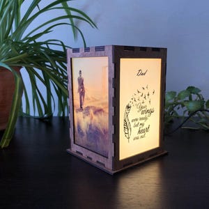 Może przedstawiać: Drewniana latarnia pamiątkowa. Z jednej strony zdjęcie osoby, z drugiej napis "Tato, Twoje skrzydła były gotowe, ale moje serce nie". Latarnia jest podświetlana od wewnątrz, tworząc delikatne światło.