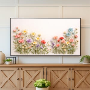 May include: A framed floral artwork featuring a variety of colorful, three-dimensional flowers. The flowers include pink, red, yellow, purple, and blue blooms, set against a white background. The artwork is displayed above a wooden cabinet.