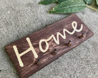 Puede incluir: Un llavero rectangular de madera teñida con la palabra "Home" en color claro. El llavero tiene cuatro pequeños ganchos de color oscuro para colgar llaves. El fondo es una superficie de hormigón con hojas verdes.