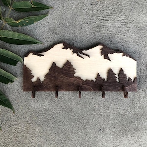 May include: Wooden key holder featuring a mountain and forest silhouette design. The key holder is made of dark brown wood with a natural wood-colored mountain and tree cutout. It has four metal hooks for hanging keys.