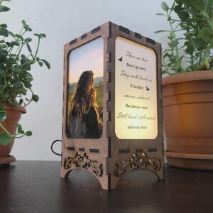 May include: A wooden memorial lamp with a photo of a person with long hair and a sunset background. The lamp has a light-up panel with the text "These we love, Don't go away, They walk beside us Everyday..." Decorative floral cutouts adorn the base.