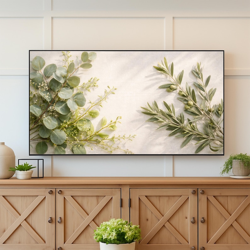 May include: A framed botanical print featuring eucalyptus and olive branches. The leaves are various shades of green, with small white flowers. The background is a light, neutral color, and the frame is black. The print is displayed on a wooden cabinet.