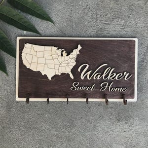 May include: Wooden key holder with a dark brown background and a light wood border. The design features a map of the United States, the name "Walker", and the phrase "Sweet Home" in white script. Includes five hooks.