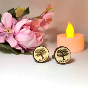 Puede incluir: Dos gemelos redondos con un diseño de árbol y la fecha 23.11.2019. Los gemelos tienen un borde de color bronce y un centro de madera clara. Una flor rosa y una vela están en el fondo.