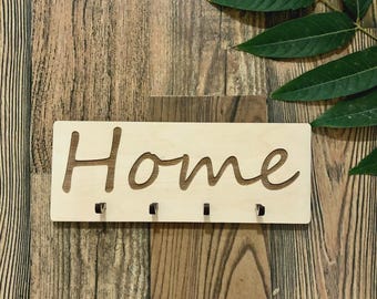 Puede incluir: Un llavero rectangular de madera clara con la palabra "Home" recortada. Tiene cuatro pequeños ganchos negros debajo. El fondo es de madera. Una hoja verde está en la esquina superior derecha.