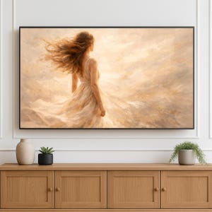 Op de afbeelding: Een ingelijste kunstdruk toont een vrouw in een vloeiende witte jurk, van achteren gezien, met haar lange, door de wind bewogen bruine haar. De achtergrond is een zacht, warm getint, abstract landschap. Het kunstwerk wordt boven een houten kast getoond.