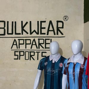 Puede incluir: Dos maniquíes exhiben camisetas deportivas. Una es de color verde azulado con rayas verde azulado oscuro, la otra es azul claro con rayas granates. Ambas tienen cuellos blancos y el texto "Webster FC". La pared detrás de ellos dice "BULKWEAR APPAREL SPORTS".