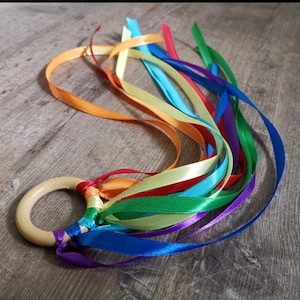 Könnte beinhalten: Ein Holzringspielzeug mit bunten Satinbändern in Rot-, Orange-, Gelb-, Grün-, Blau- und Lilatönen. Die Bänder sind an dem Ring befestigt und bilden ein sensorisches Spielzeug für Babys und Kleinkinder.